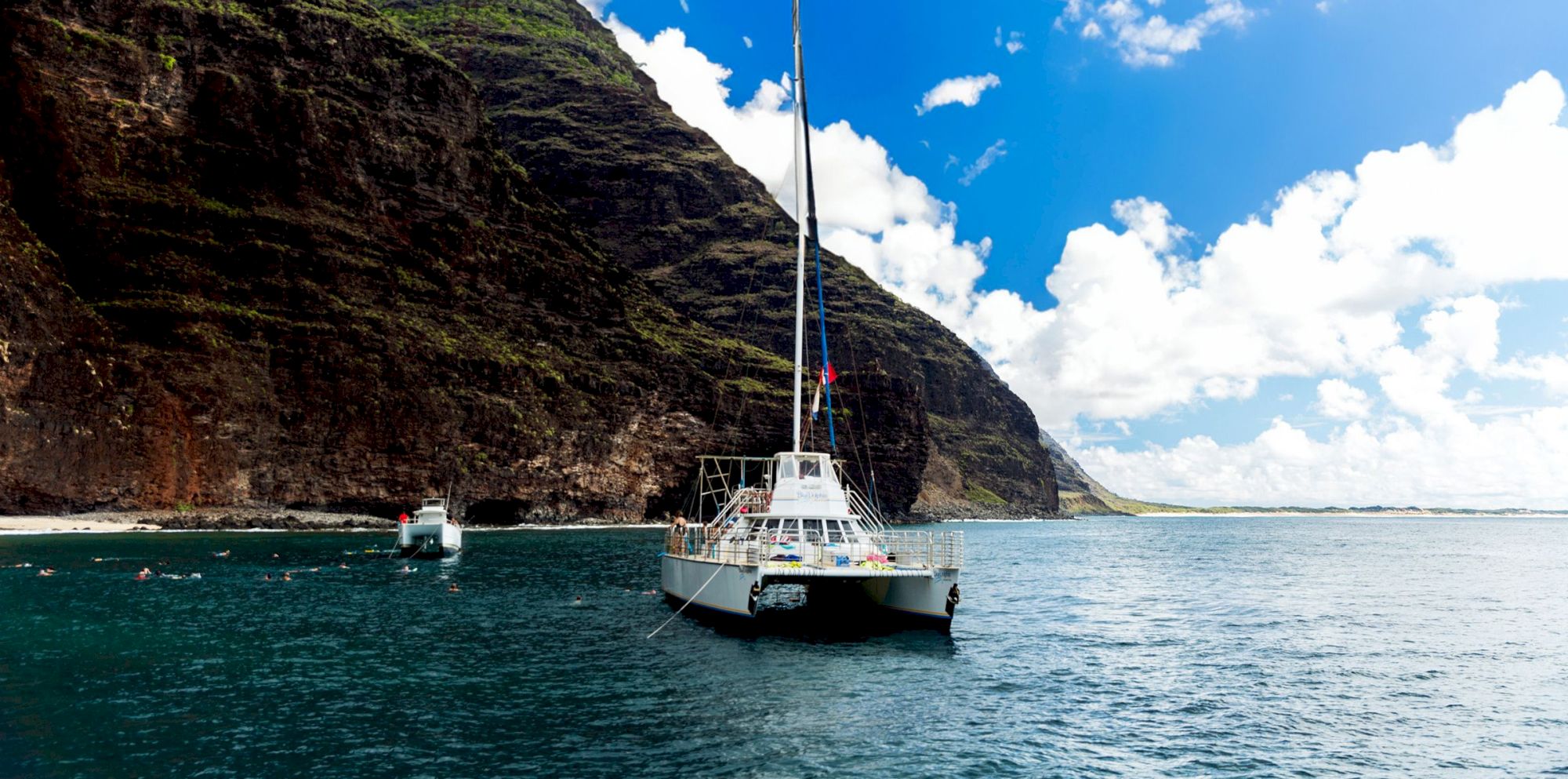 A catamaran is sailing near a rocky coastal area with lush greenery, under a bright blue sky with scattered clouds.