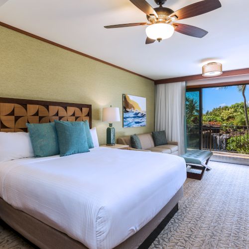 A spacious hotel room features a king-size bed, ceiling fan, and a balcony with a vibrant garden view.