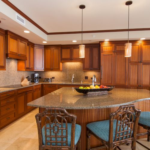 A modern kitchen with wooden cabinetry, a large island, granite countertops, barstools, pendant lights, and stainless steel appliances.