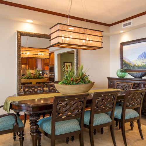 The image shows a dining room with a wooden table, six chairs, a large plant centerpiece, a mirror, and a mountain landscape painting.