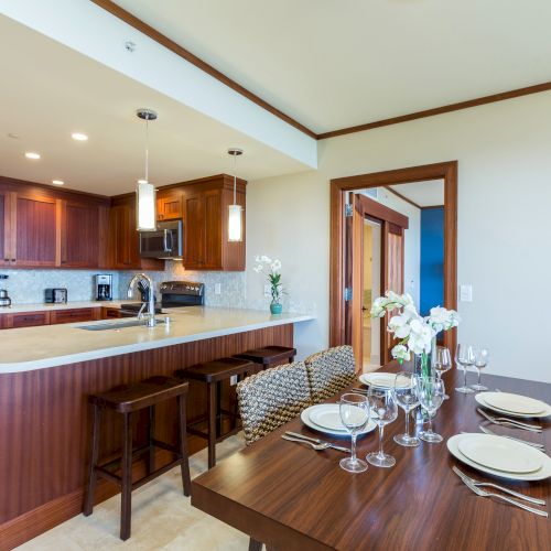 A modern kitchen and dining area with wooden cabinets, a countertop, bar stools, and a dining table set with plates and glasses.