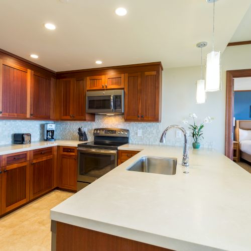 A modern kitchen with wooden cabinets, stainless steel appliances, and a white countertop; a bedroom is visible in the background.