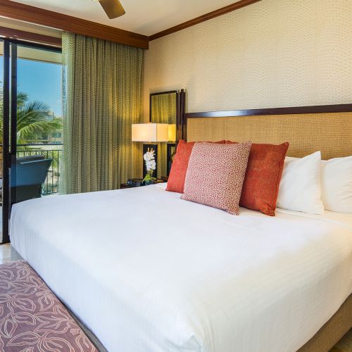 A hotel room with a large bed, orange pillows, lamps, and a balcony with a view of palm trees and outdoor seating.