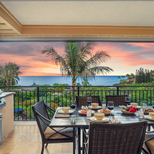 A patio dining setup overlooks a scenic ocean view at sunset, featuring a grill and a table set with drinks and dinnerware.