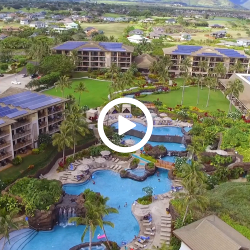 Aerial view of a resort featuring pools, landscaped gardens, and buildings with solar panels surrounded by scenic greenery and mountains.