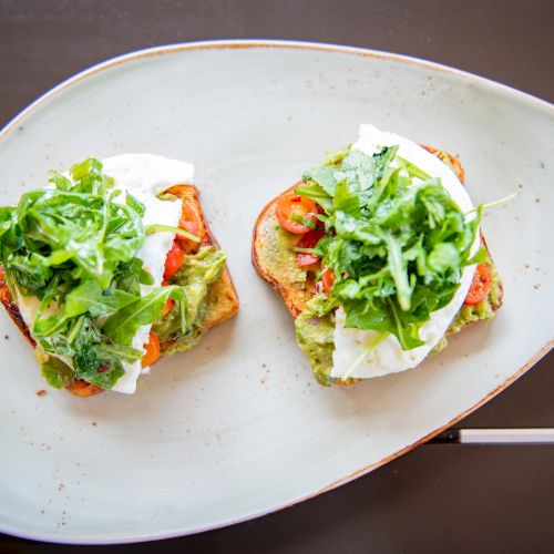 Two slices of toast topped with avocado, arugula, tomato, and mozzarella are arranged on a stylish plate for serving.
