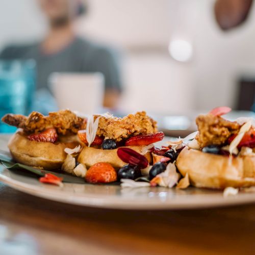 A plate with waffles topped with fried chicken, berries, and sauce, is on a table. People are blurred in the background.