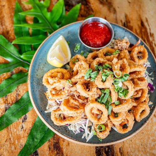 A plate of fried calamari with lemon, green garnish, and a side of red dipping sauce, placed on a wooden surface with green leaves.