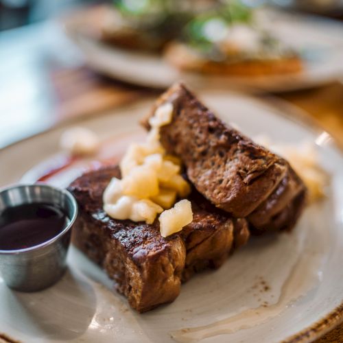 Plate with French toast topped with diced apples and syrup on the side, served on a wooden table.