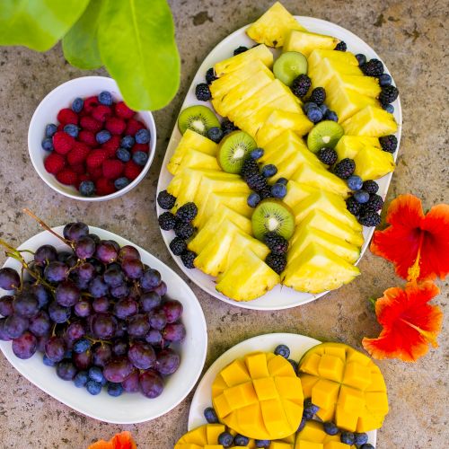 The image shows plates of sliced pineapple, mango, kiwi, blueberries, raspberries, and grapes on a stone surface with vibrant orange flowers.