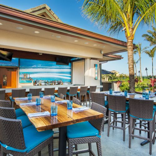 Outdoor dining area with tables, chairs, a mural, and palm trees under a clear blue sky.