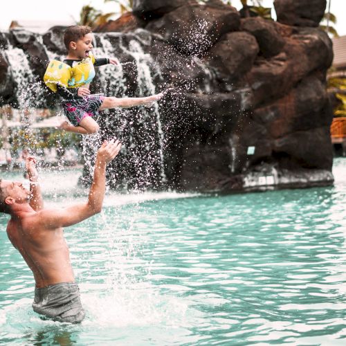A person is playfully tossing a child wearing a life jacket into the air above a pool with a waterfall feature.