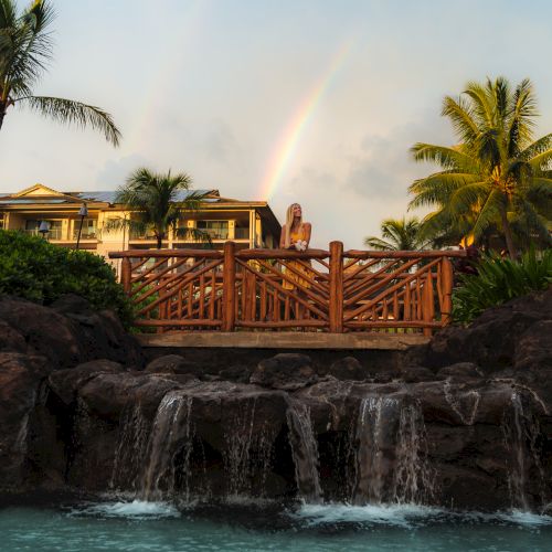 A tropical scene features a wooden bridge, a small waterfall, lush palm trees, and a rainbow in the background under a cloudy sky.