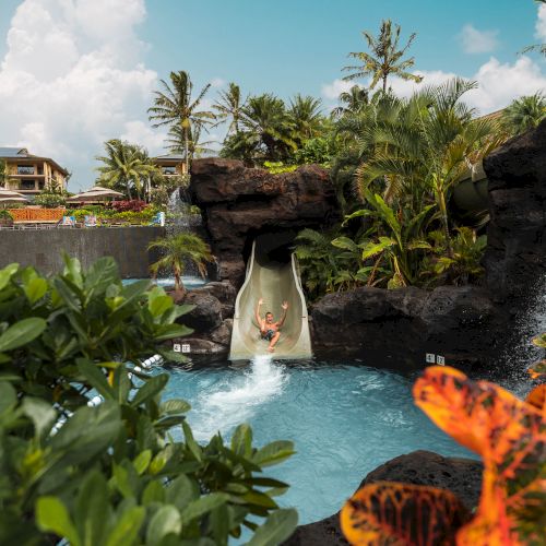 A person slides down a water slide into a pool surrounded by lush tropical plants and trees under a clear blue sky.