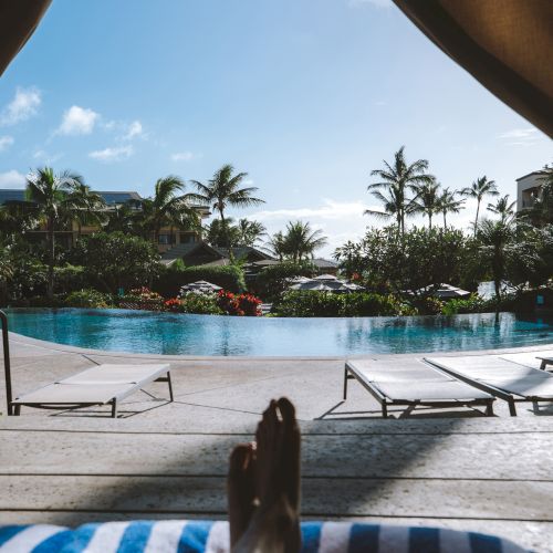 A pair of feet rests poolside on a blue-striped towel, with a view of palm trees and blue skies beyond the shimmering water.