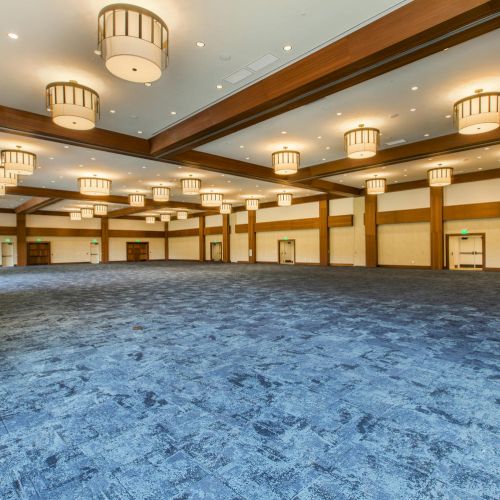 A large, empty banquet hall with blue carpet and multiple ceiling lights and wooden beams is shown in the image.