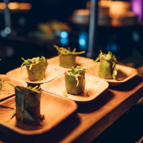 Plates with neatly arranged rolls of greens, possibly hors d'oeuvres, are displayed on a wooden table under warm lighting.