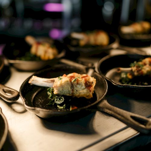 Several small skillets with plated gourmet dishes featuring a piece of meat and garnish, arranged on a counter in a dimly lit environment.