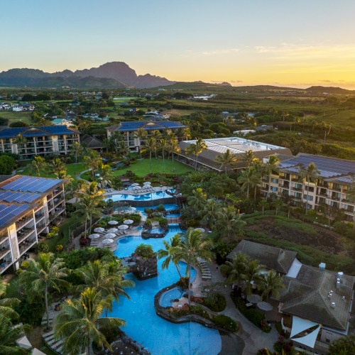 Aerial view of a resort with pools, surrounded by palm trees and buildings at sunset, with a mountain in the background.