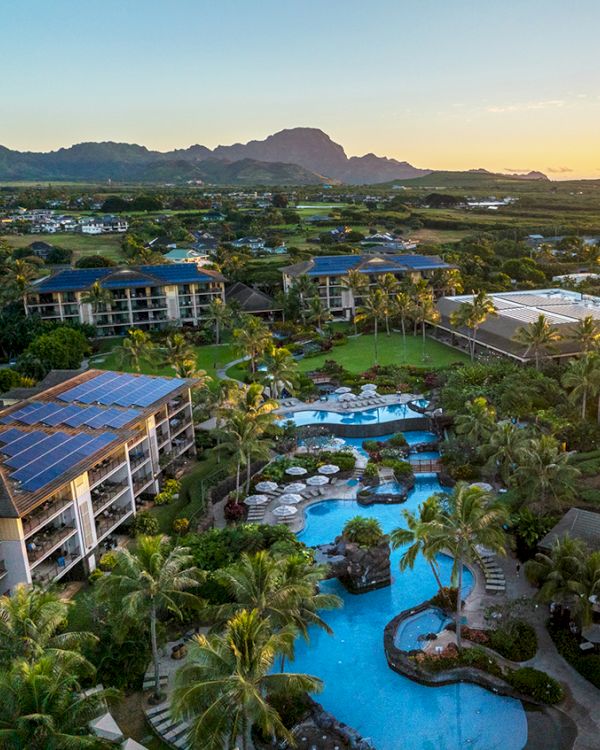 Aerial view of a resort with pools, palm trees, and buildings, surrounded by lush greenery and mountains.