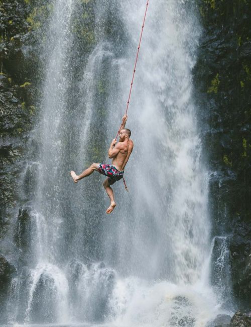 A person is swinging on a rope in front of a waterfall, surrounded by rocks and greenery.
