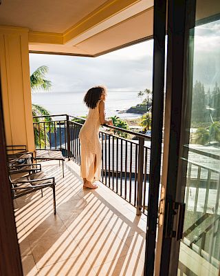 A person in a dress stands on a balcony, gazing at the ocean view, with tropical surroundings and sunlight casting shadows.