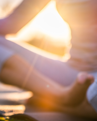A person is meditating outdoors in the lotus position on a yoga mat, with sunlight shining through in the background, focusing on relaxation.